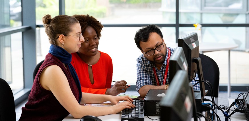 Two female students sit around a computer with a male student.